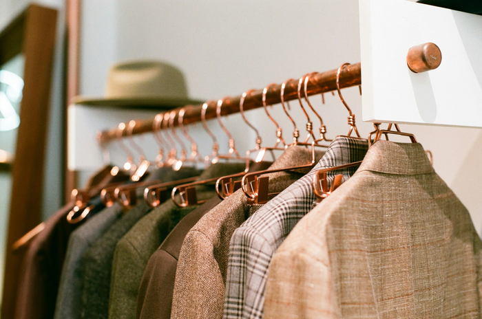 a rail of clothes. The rail is a rose gold colour, with rose gold hangers, with blazers attached to them. There is a hat in the top left corner.